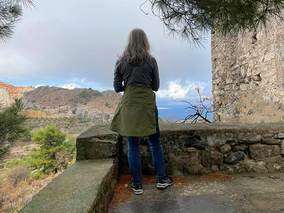 Valeria stands with her back to the camera facing a landscape of old stone walls, dry hills, and scrub brushes. Her hair is gray and loose around her shoulders and she is dressed in comfortable clothes of jeans, a gray shirt, and a green coat tied around her waist with black sneakers.